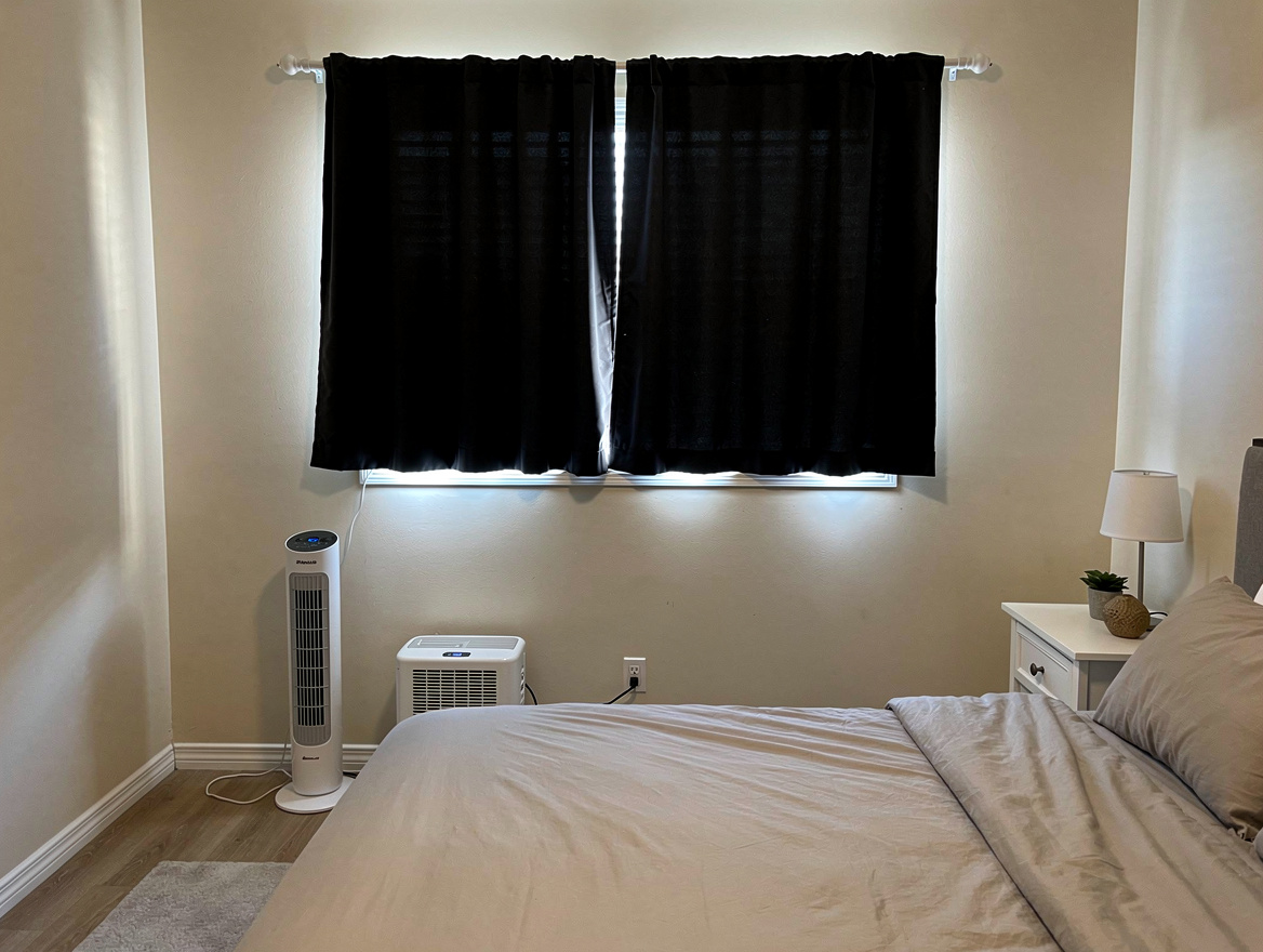 Medium shot of a bedroom with blackout curtains on a tension rod, slim tower fan